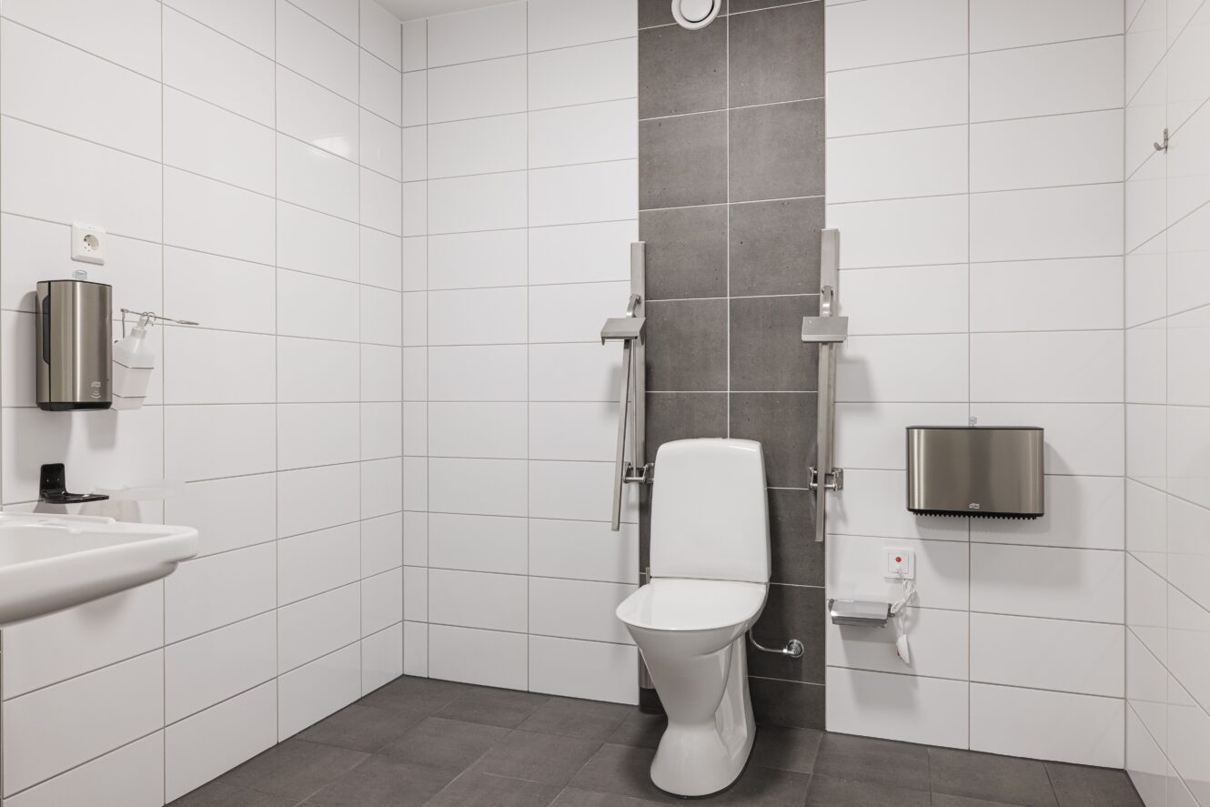 Tillgänglig offentlig toalett med vitkaklade väggar, toalett med stödskenor, handfat, tvålpump, pappershanddukspump och nödstopp.