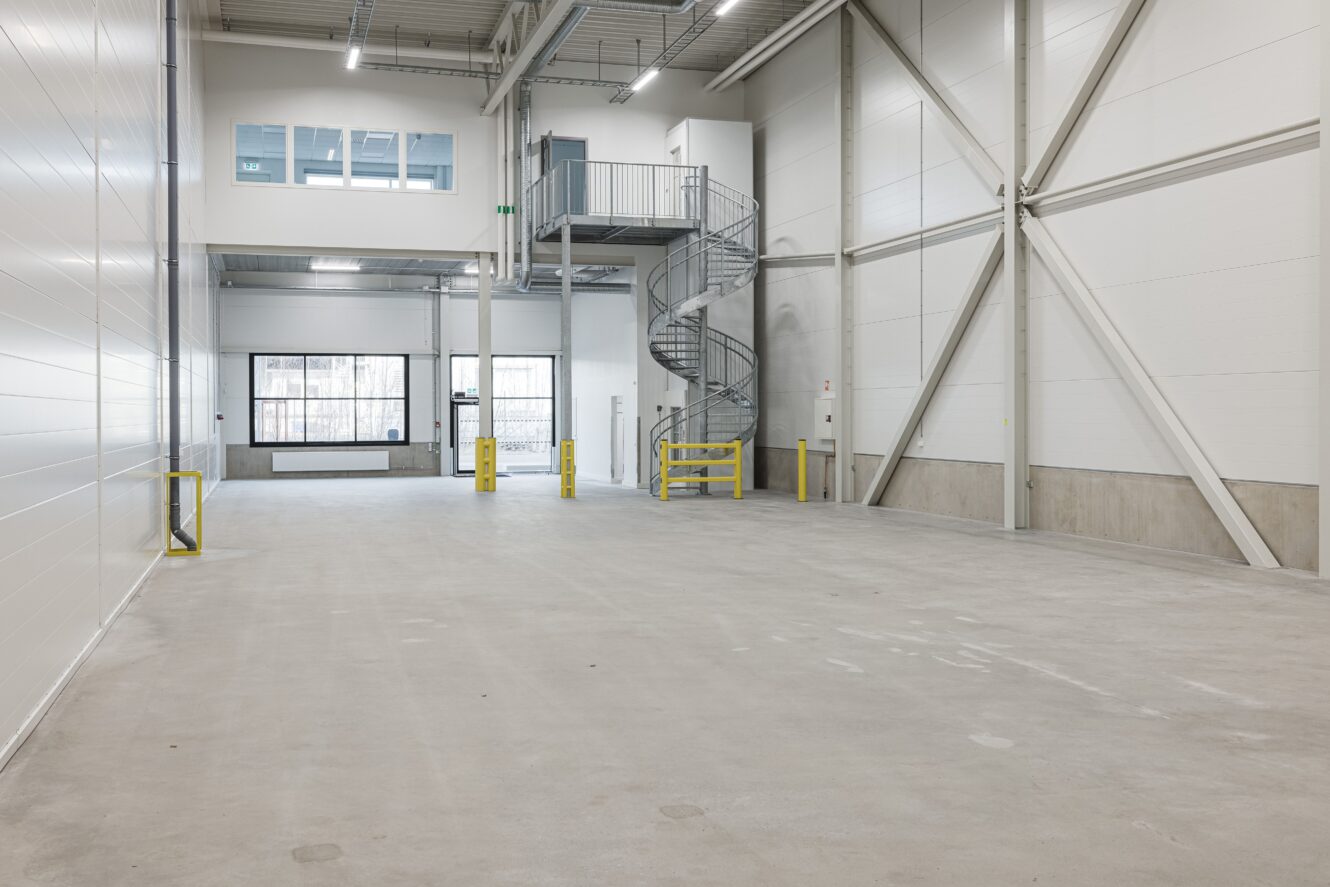 Tomt industrilager med betonggolv, vita väggar, stora fönster, gula säkerhetsbarriärer och en spiraltrappa i metall som leder till en övre våning.