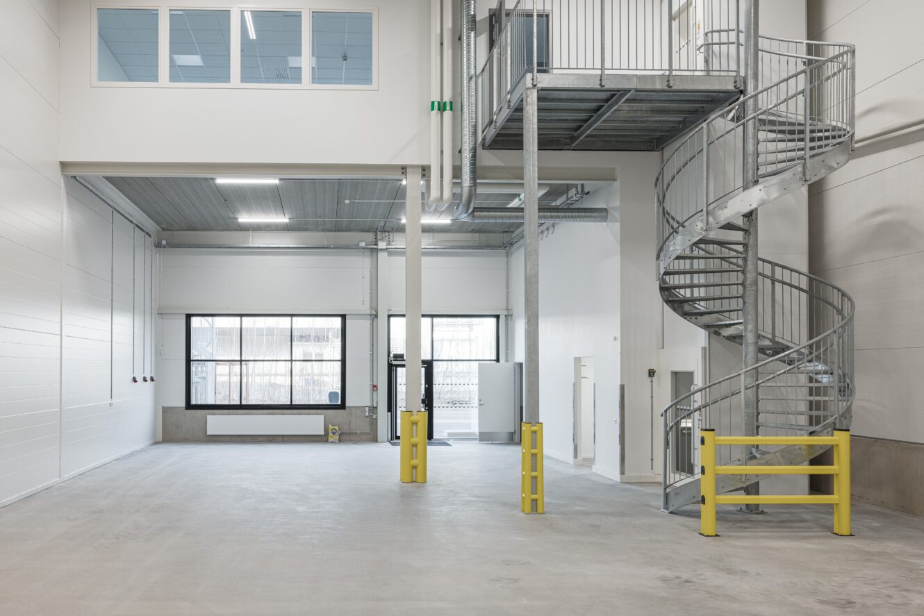Rymlig, tom industriinredning med vita väggar, stora fönster och en spiraltrappa i metall som leder till en mezzaninvåning; gula säkerhetsbarriärer finns.