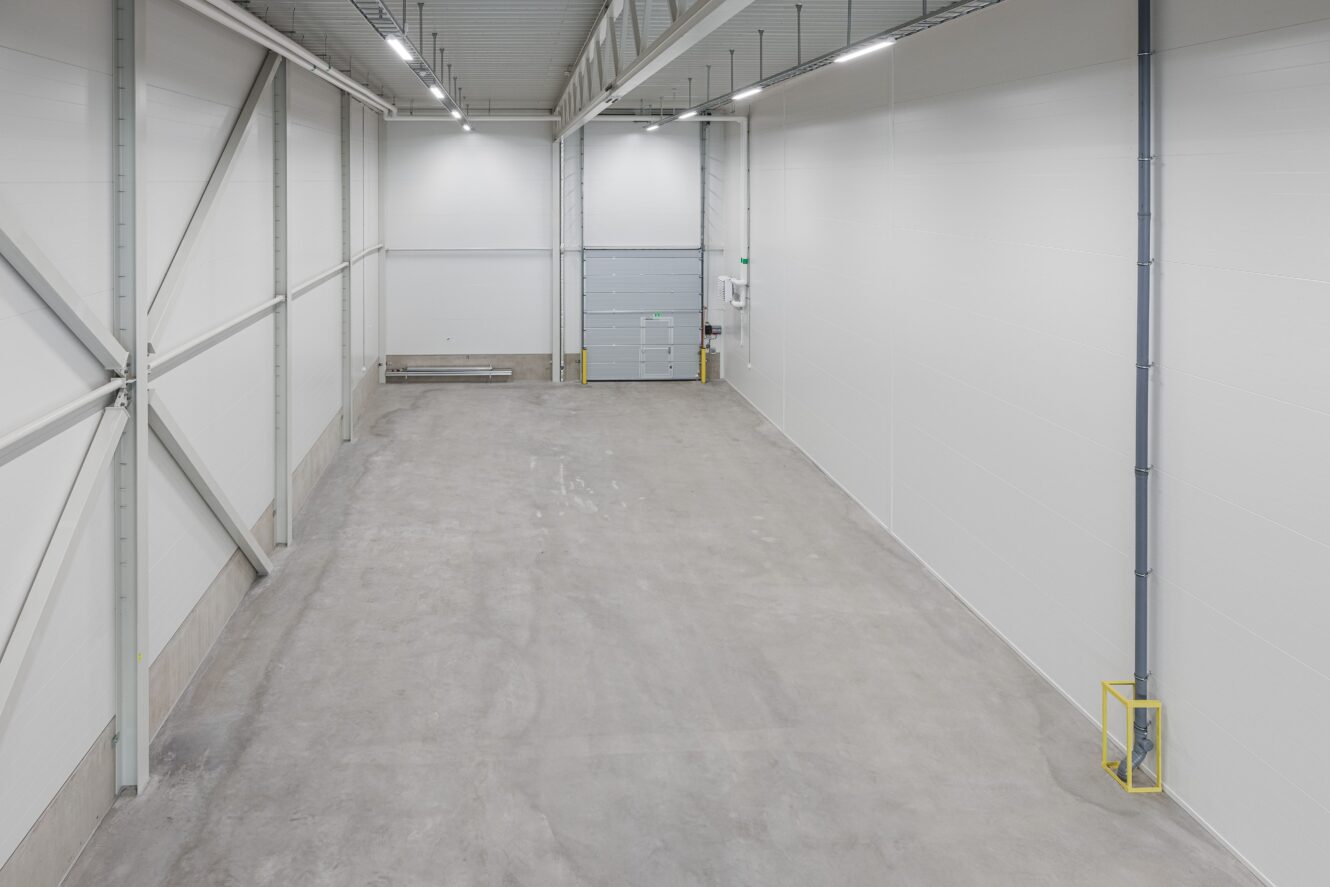 Stort tomt industriellt lagerutrymme med vita väggar, synliga rörledningar, högt i tak och en stängd garageport längst bort.