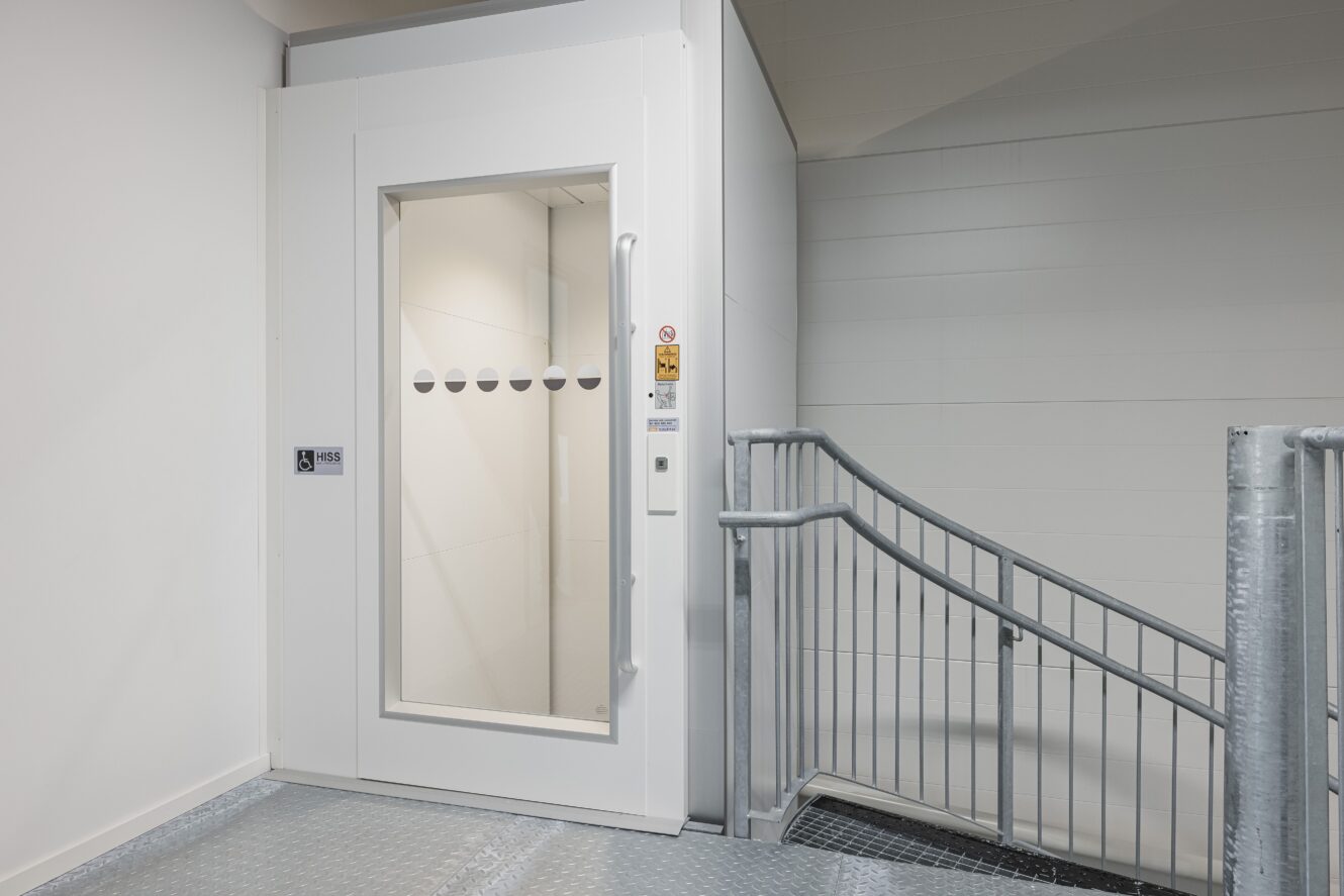 En modern, vit hiss med glasdörr är installerad bredvid en metalltrappa i en inomhusmiljö.
