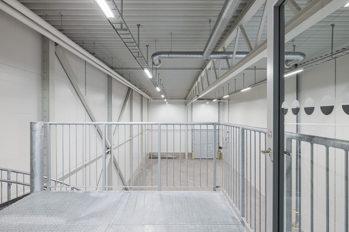 Tomt industriellt lagerutrymme med högt i tak, metallräcken, synliga kanaler och en stor garageport, sett från en mezzaninvåning.