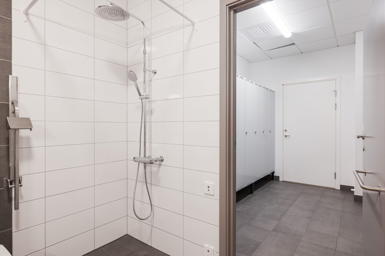 Ett rent omklädningsrum med vita kaklade väggar har ett duschutrymme och en rad vita skåp och bås, med gråa klinkergolv och takbelysning.