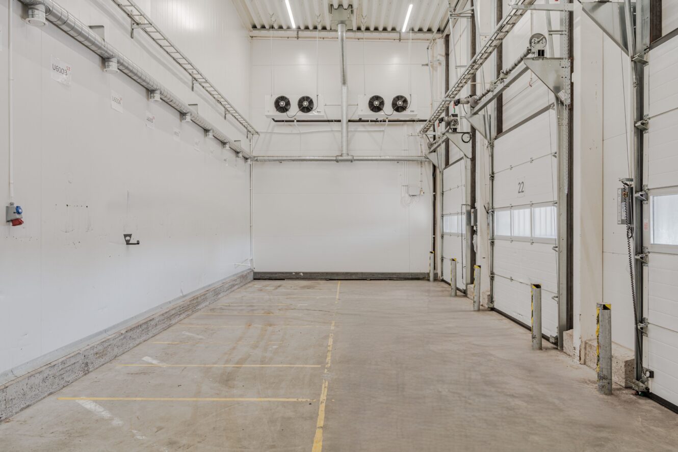 Interiör i en tom lagerlokal med vita väggar, takbelysning, tre lastbryggor och ventilationsfläktar på bakre väggen.