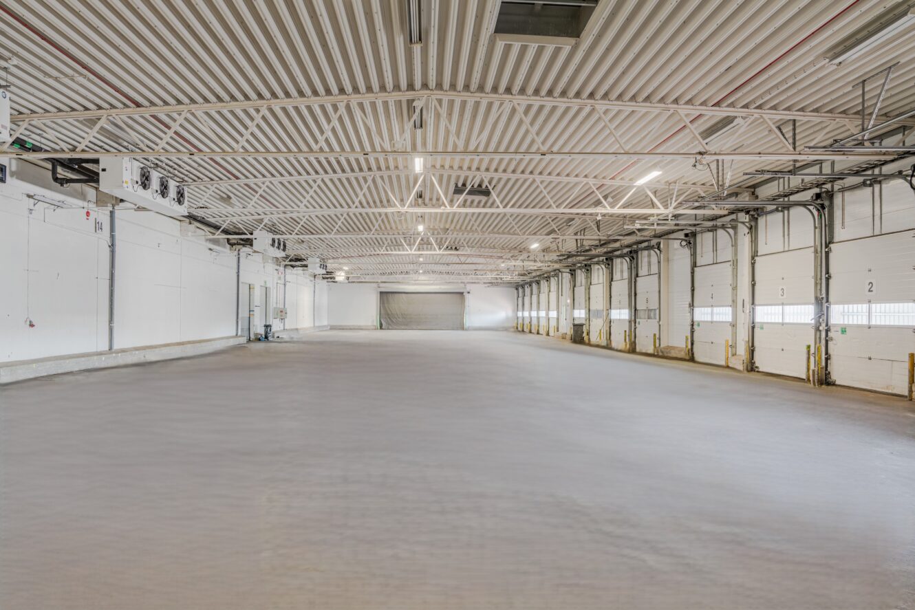 Interiör av ett stort, tomt industrilager med högt i tak, vita väggar och flera lastkajsdörrar längs ena sidan.