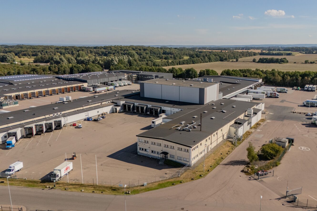 Flygfoto över ett stort industriellt lagerkomplex med flera lastbryggor, parkerade lastbilar och omgivande grönska en klar dag.