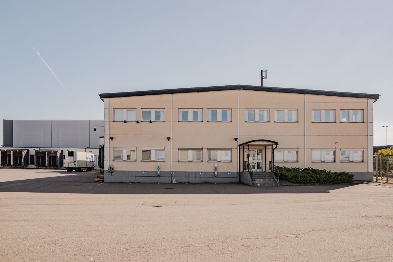 En tvåvånings industribyggnad med flera fönster, en central entré med trappor och en lastbil parkerad vid lastbryggor till vänster.