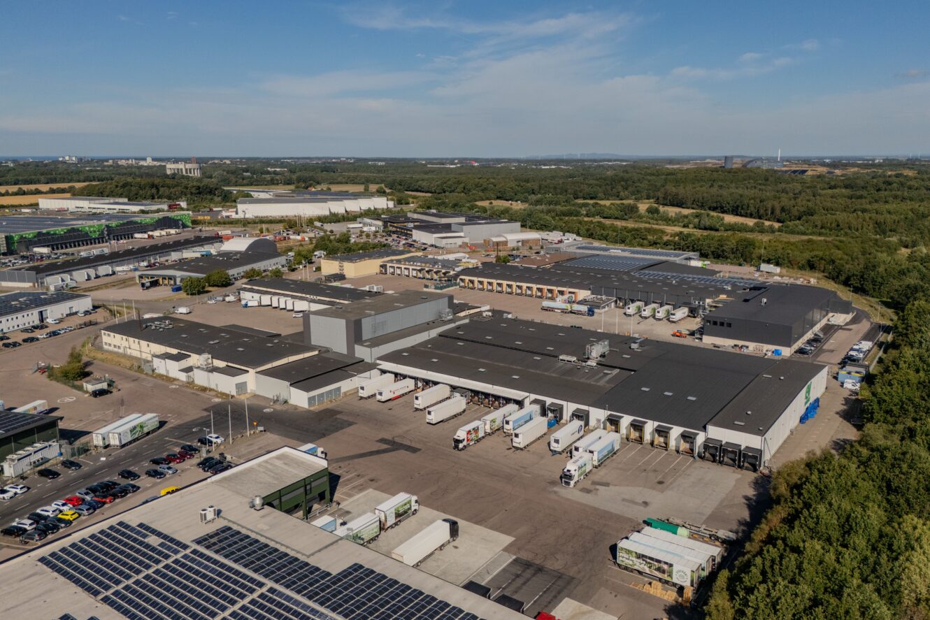 Flygfoto över ett stort industrikomplex med flera lagerlokaler, parkerade lastbilar och solpaneler på vissa tak, omgivet av grönska under en klar himmel.