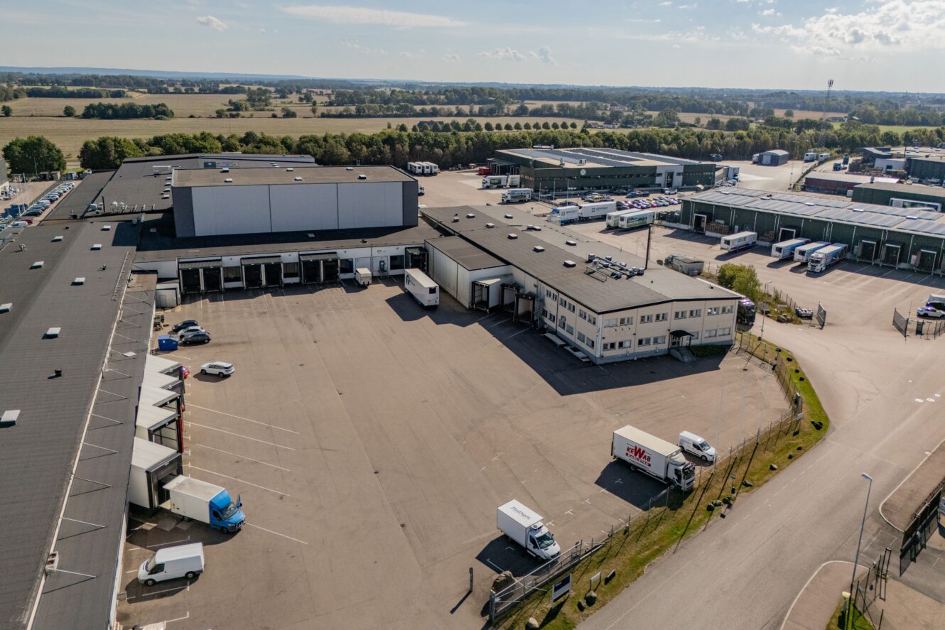 Flygfoto över en logistikanläggning med lagerlokaler, parkerade lastbilar, lastkajer och en intilliggande parkeringsplats en klar dag.