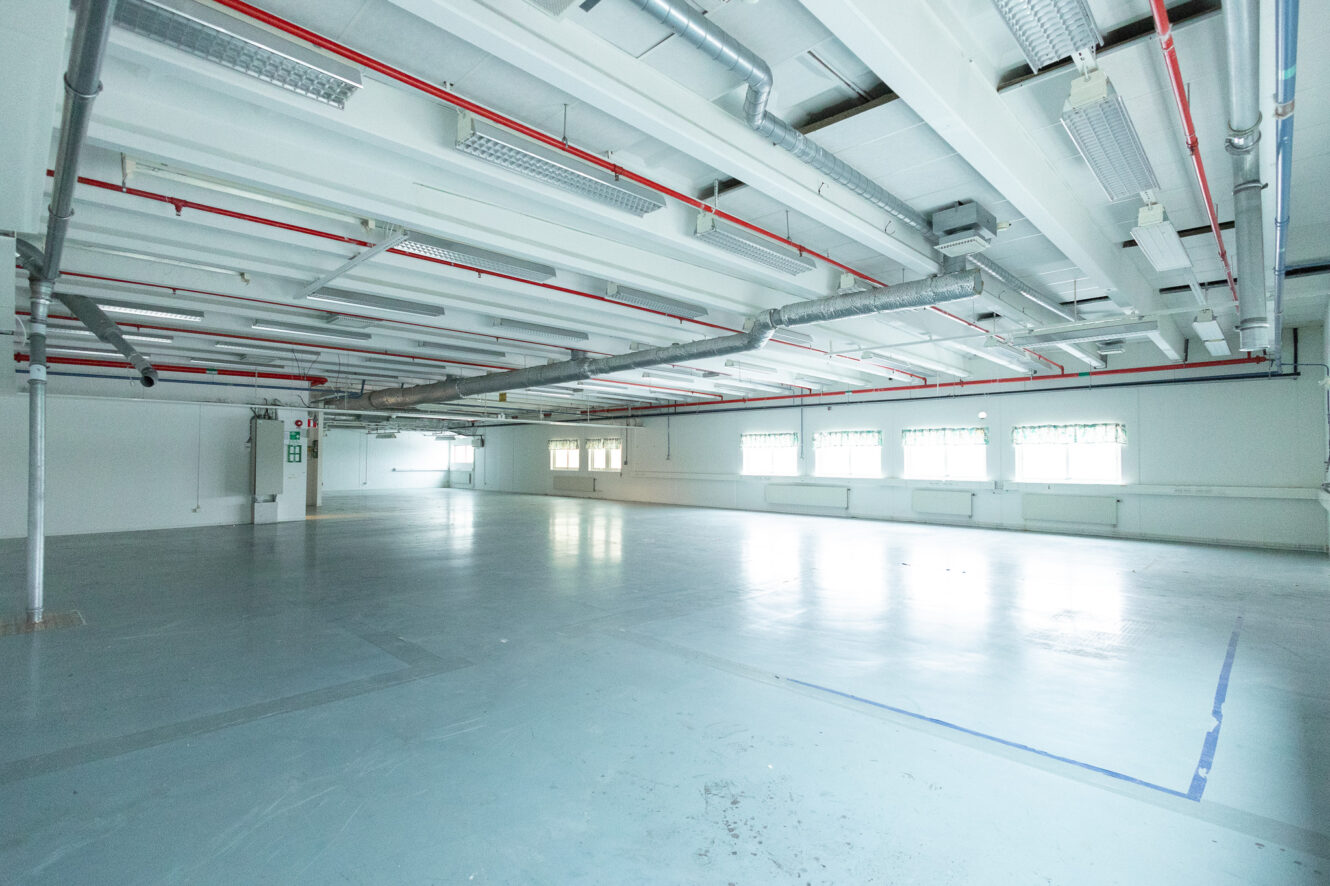 Stort tomt industrirum med vita väggar, synliga rör i taket, lysrör i taket och flera fönster som släpper in naturligt ljus.