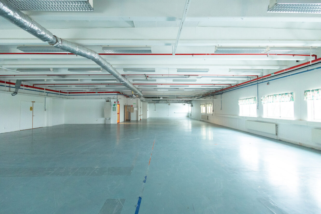 Stort tomt industri- eller affärsrum med vita väggar, synliga rör i taket, blågrått golv och fönster med mönstrade persienner på höger sida.