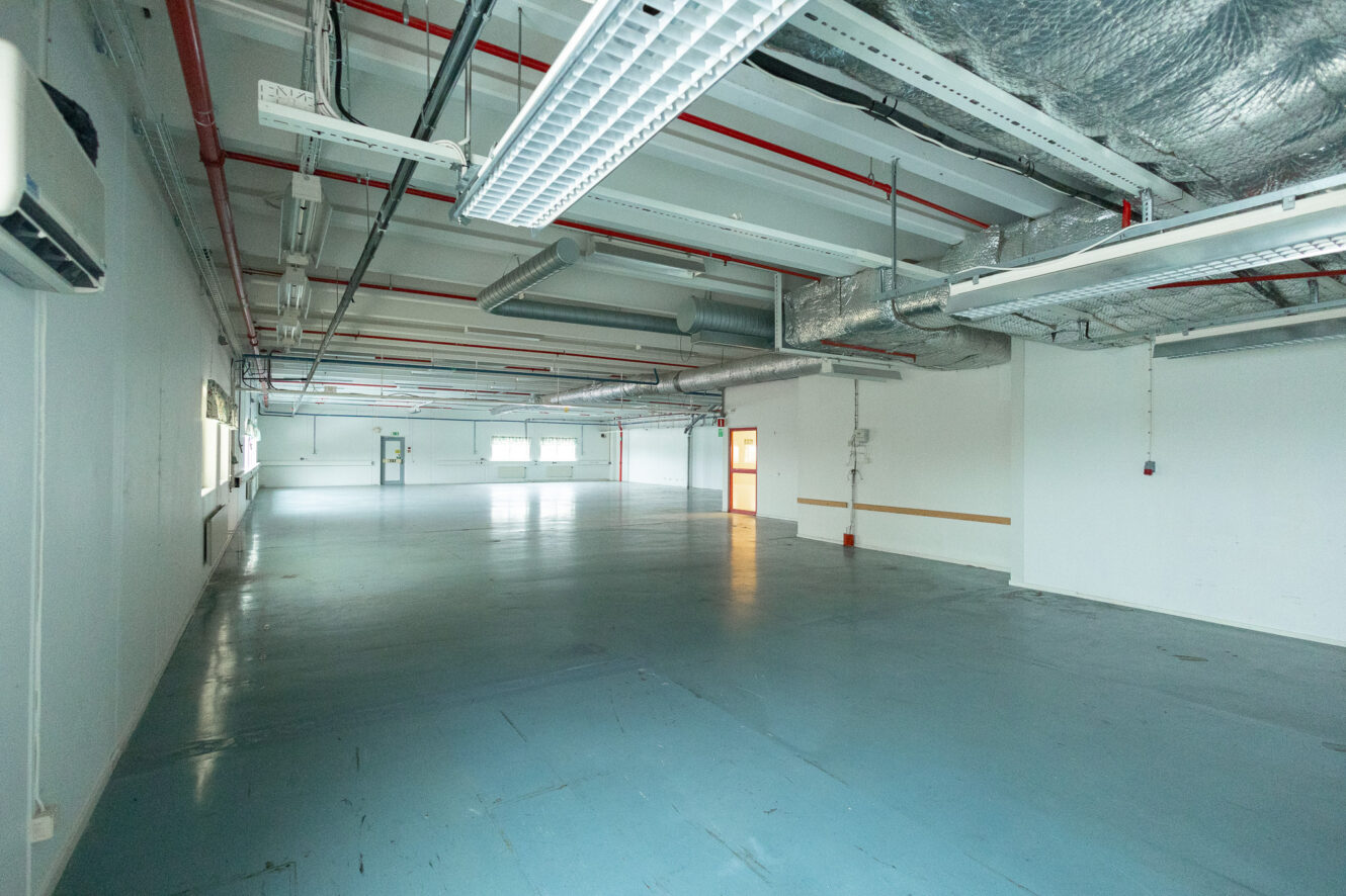 Stort tomt industrirum med blått golv, synliga rörledningar och lysrör i taket; ljus strömmar in från fönster på bortre väggen.