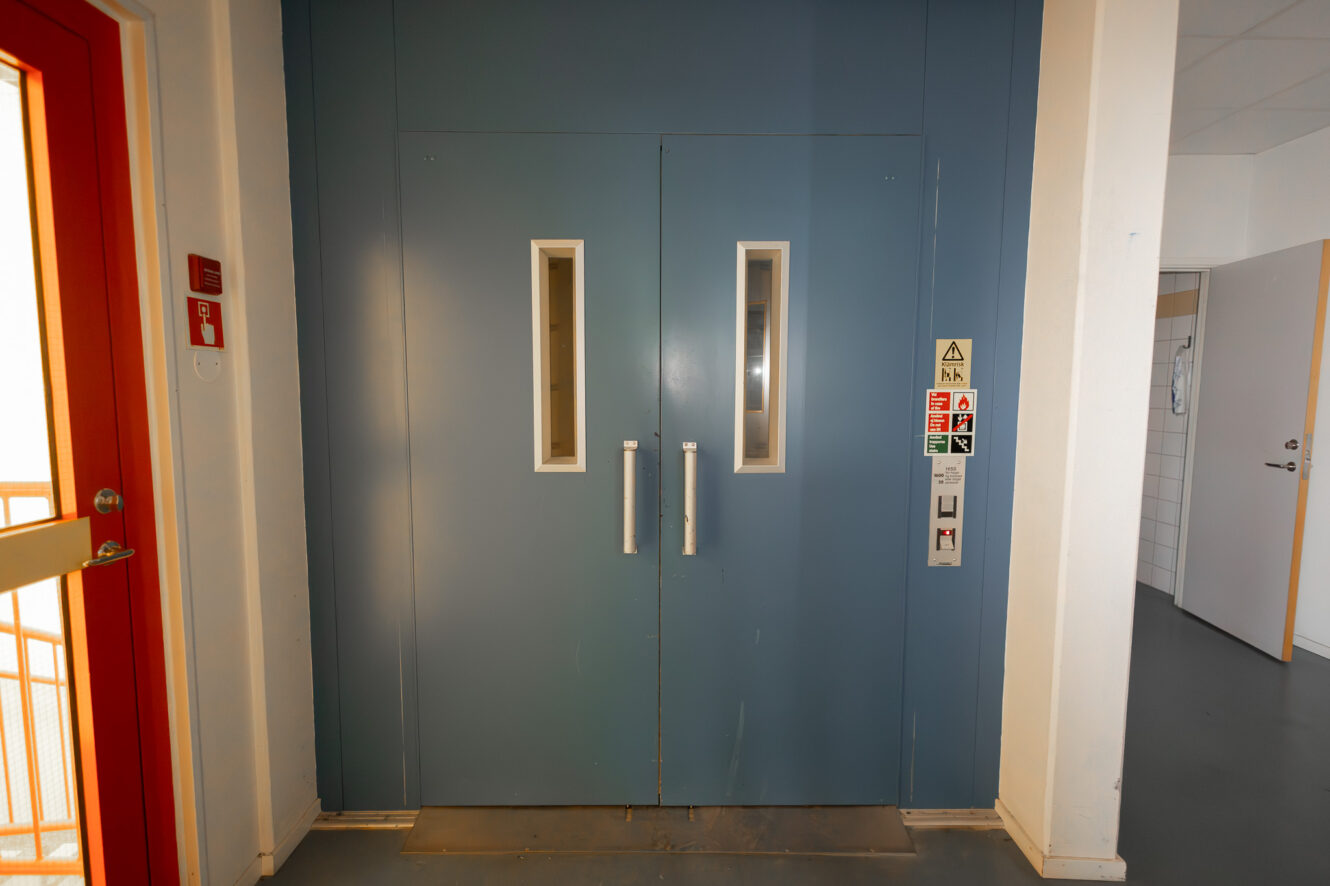 Stängda dubbla blå hissdörrar med rektangulära fönster och metallhandtag, manöverknappar och säkerhetsskyltar synliga på höger sida.