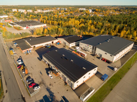 Flygfoto över ett industriområde med flera stora lagerbyggnader, parkerade bilar och träd med höstlöv i bakgrunden.