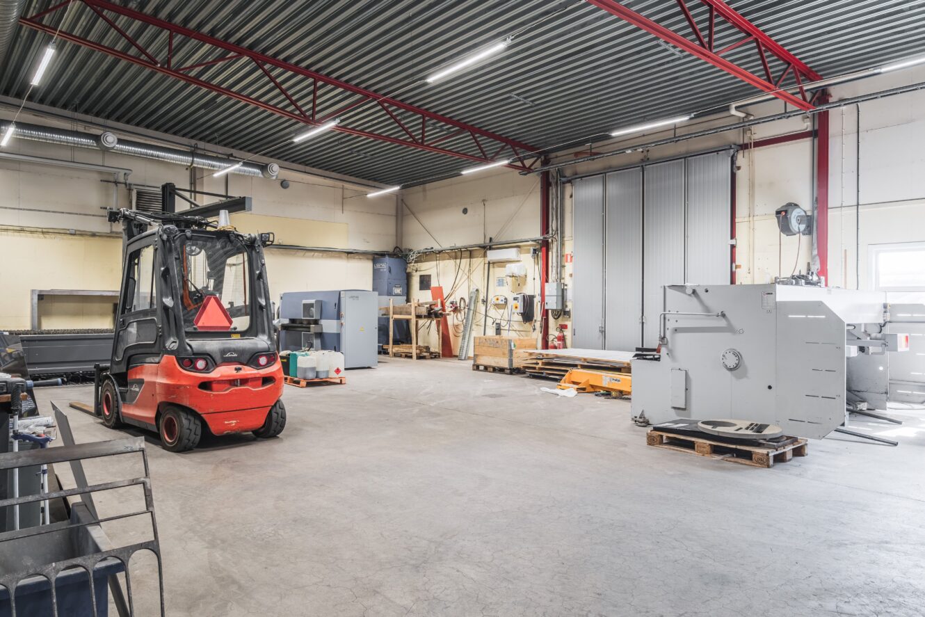 Ett rymligt industrilager med en röd gaffeltruck, olika maskiner, pallar och utrustning på ett betonggolv under fluorescerande belysning.