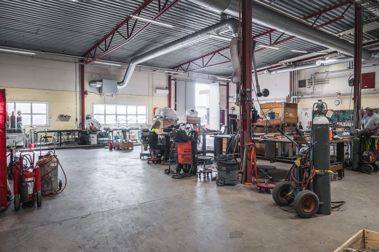 Rymlig industriverkstad med utspridd utrustning, gasflaskor, verktyg, brandsläckare och arbetsstationer; en person arbetar i bakgrunden.