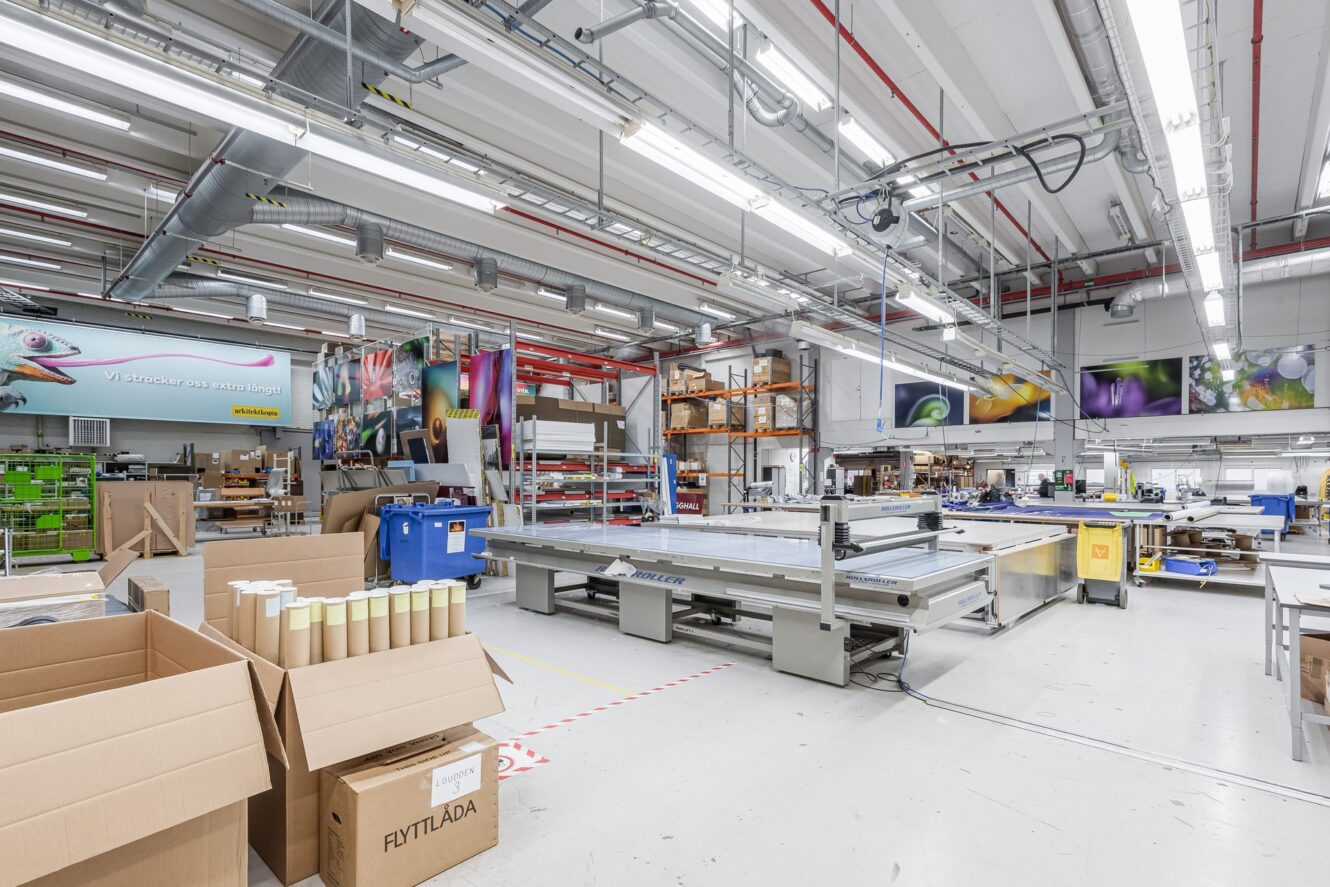 En rymlig, väl upplyst tryckeriverkstad med stora maskiner, kartonger, materialrullar och färgglada affischer på väggarna.