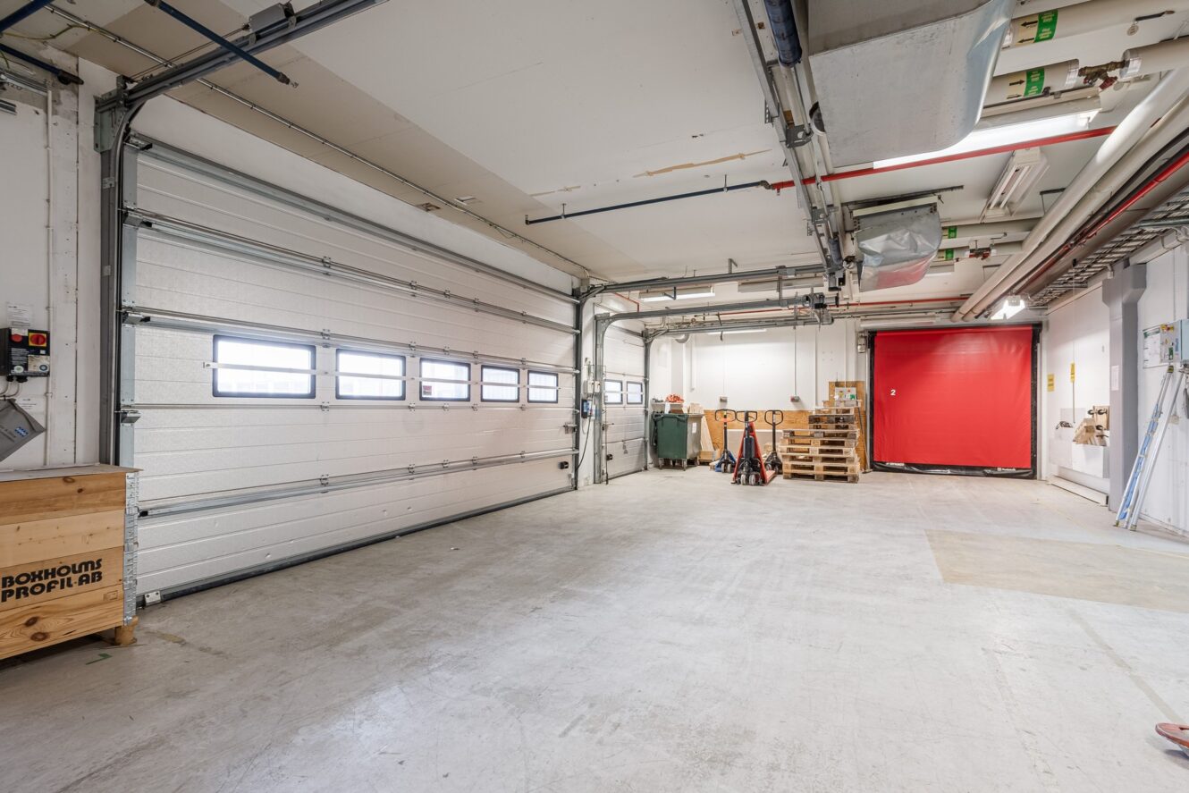 Rymligt industrigarage med en stor vit takskjutport, röd rullport, träpallar och utrustning längs väggarna.