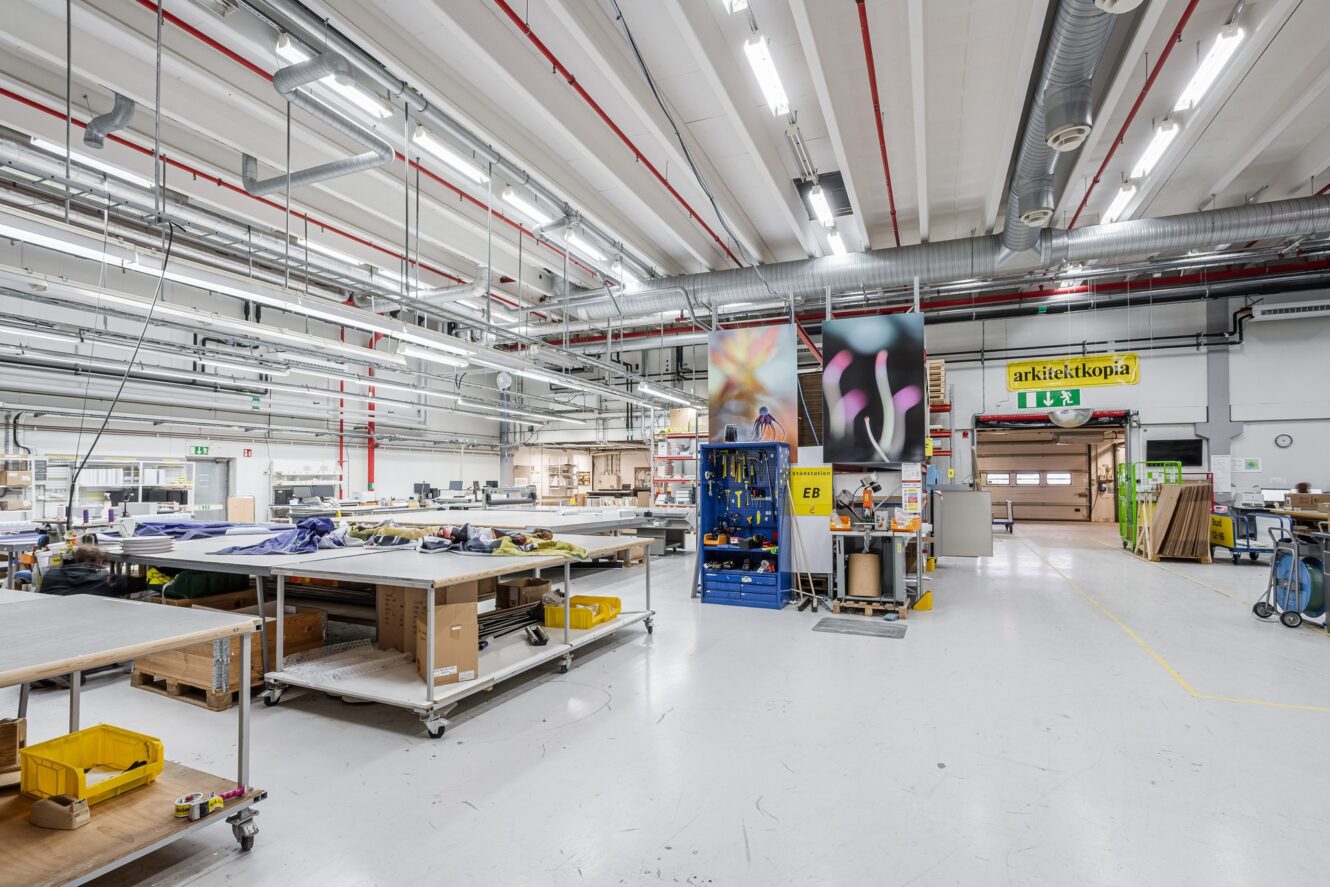 Rymlig industriverkstad med arbetsbänkar, förvaringshyllor, utrustning och takbelysning; förnödenheter organiserade över hela området.