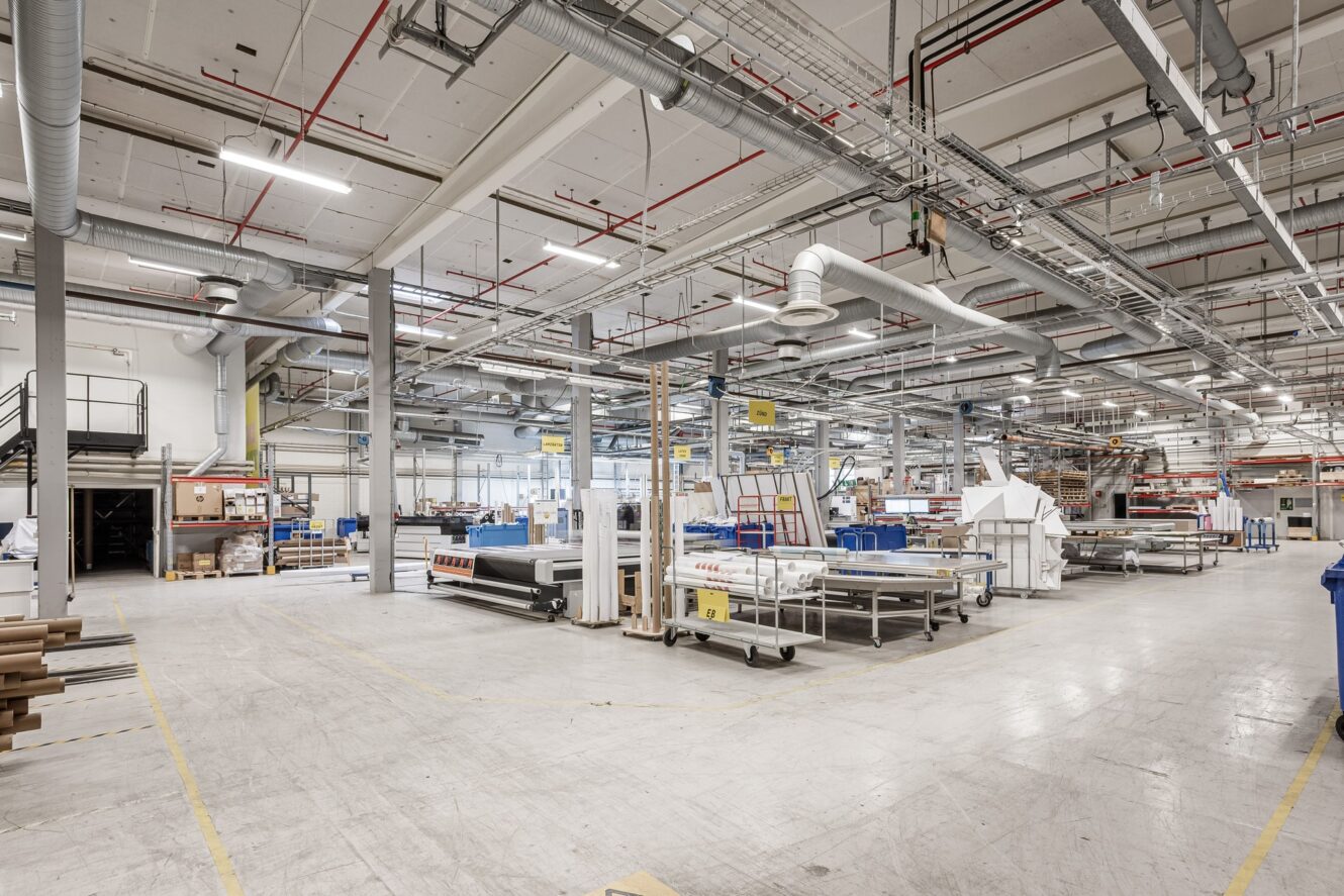 Rymligt industriellt fabriksgolv med maskiner, arbetsbord, förvaringshyllor, organiserad utrustning och starka taklampor.