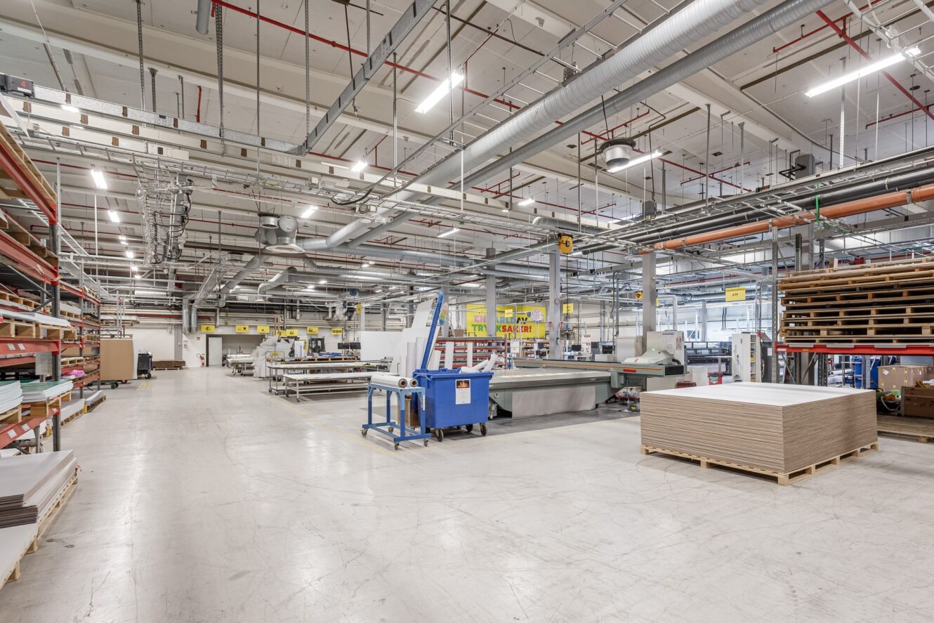 En rymlig, organiserad fabriksinredning med industrimaskiner, staplade material, metallhyllor och synliga rör i taket. Väl upplyst.