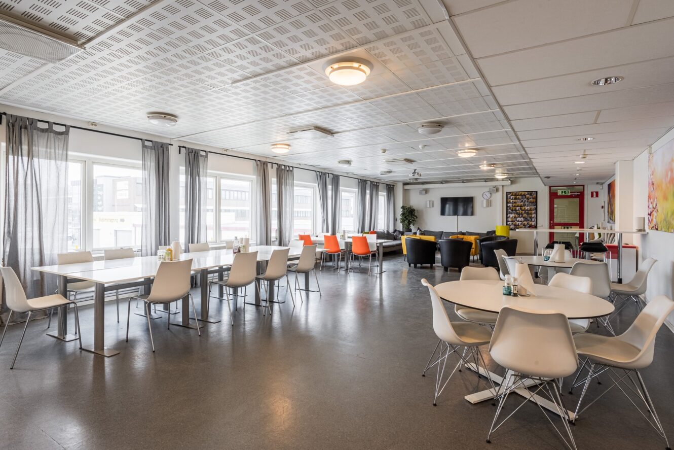 Cafeteria med vita bord och stolar, orange och svarta loungemöbler, stora fönster och en TV monterad på bakväggen.