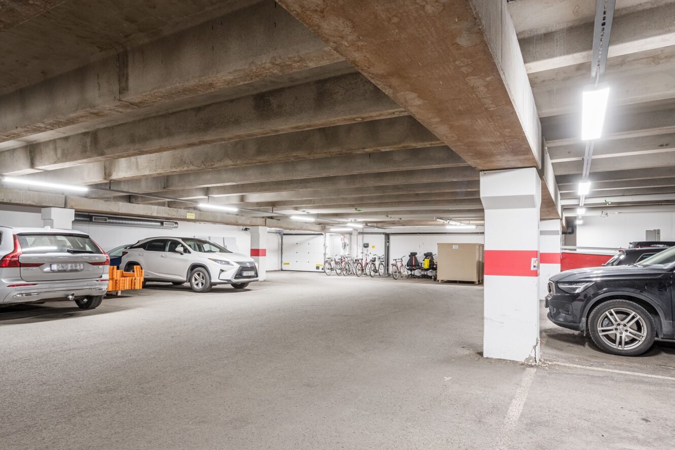 Underjordiskt parkeringsgarage med parkerade bilar, lysrörsbelysning, betongtak och cyklar uppradade längs bakväggen.