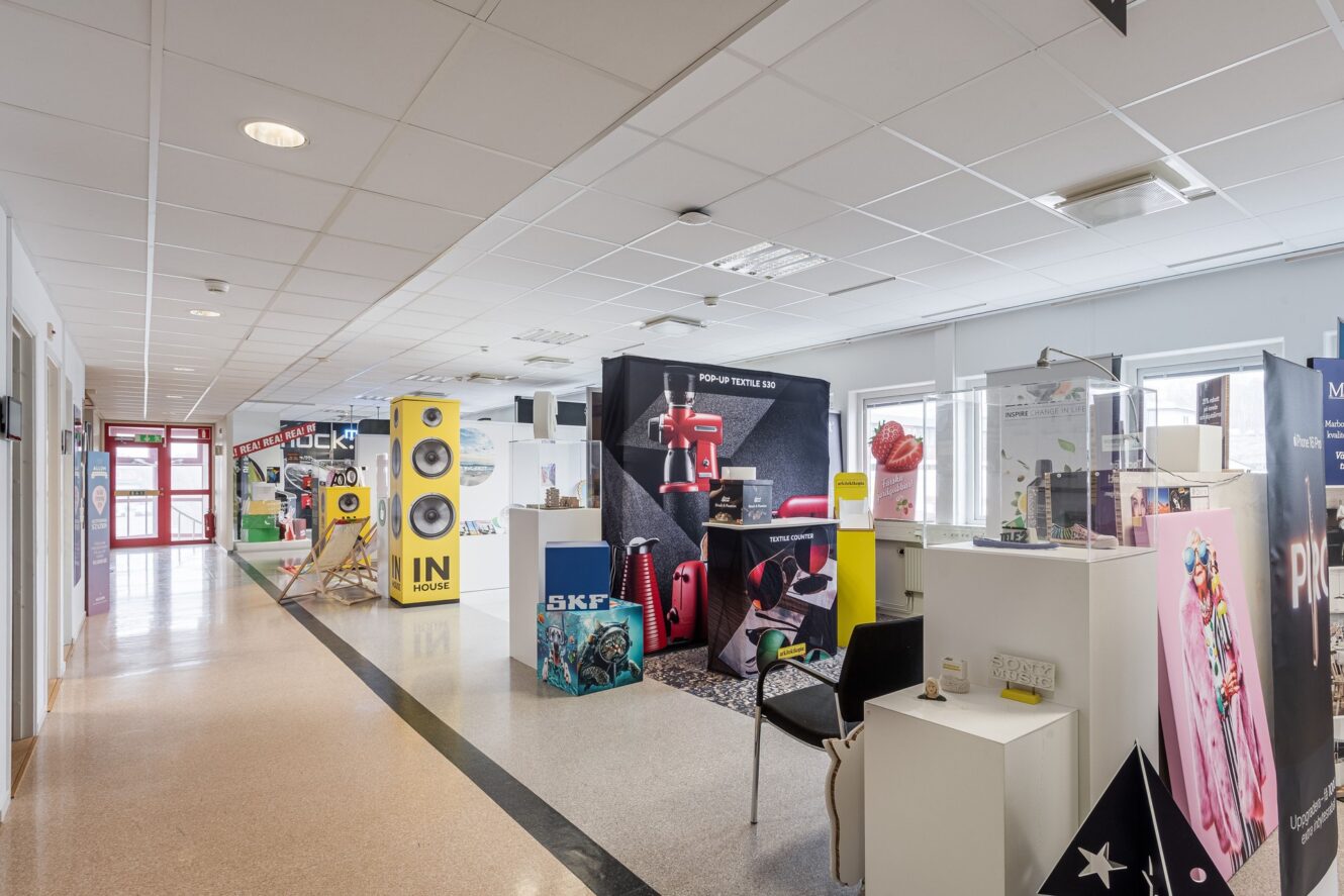 En korridor med färgglada montrar och utställningsföremål på ena sidan, under fluorescerande ljus, med fönster på den motsatta väggen.