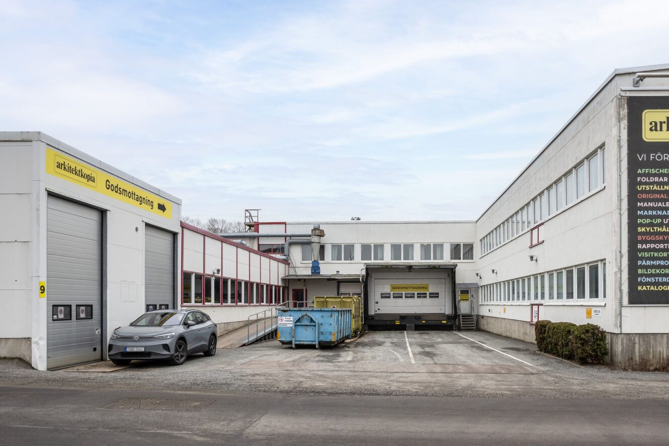 En industribyggnad med en lastkaj, en blå container, en parkerad bil och textskyltar på de vita ytterväggarna.