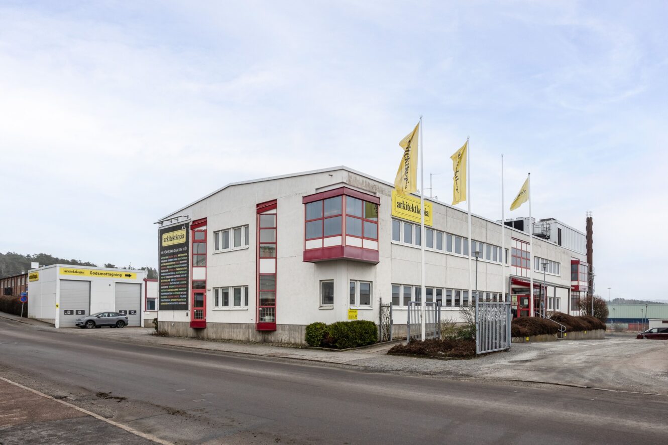 Stor vit industribyggnad med rödmålade fönster, gula skyltar och flaggor på en asfalterad gata under en molnig himmel.