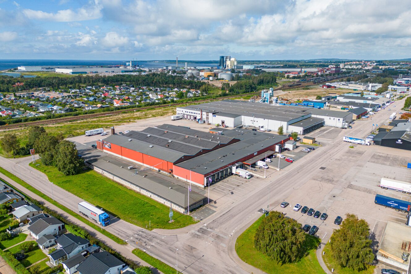 Flygfoto över ett stort industriellt lagerkomplex omgivet av vägar, parkerade lastbilar och närliggande bostadsområden med en avlägsen kustlinje.