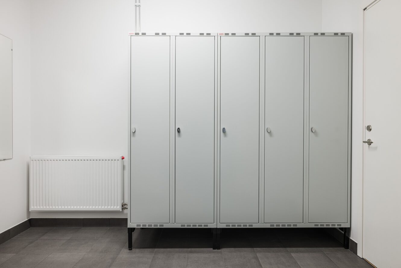 En rad med fyra stängda grå metallskåp står mot en vit vägg i ett rent och tomt omklädningsrum.