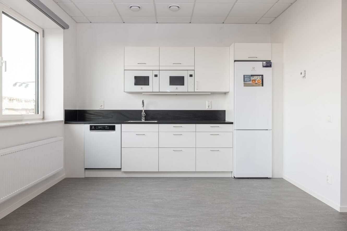 Ett modernt, minimalistiskt kök med vita skåp, en inbyggd diskmaskin, diskho, kylskåp och mikrovågsugn, mot ett ljusgrått golv och vita väggar.