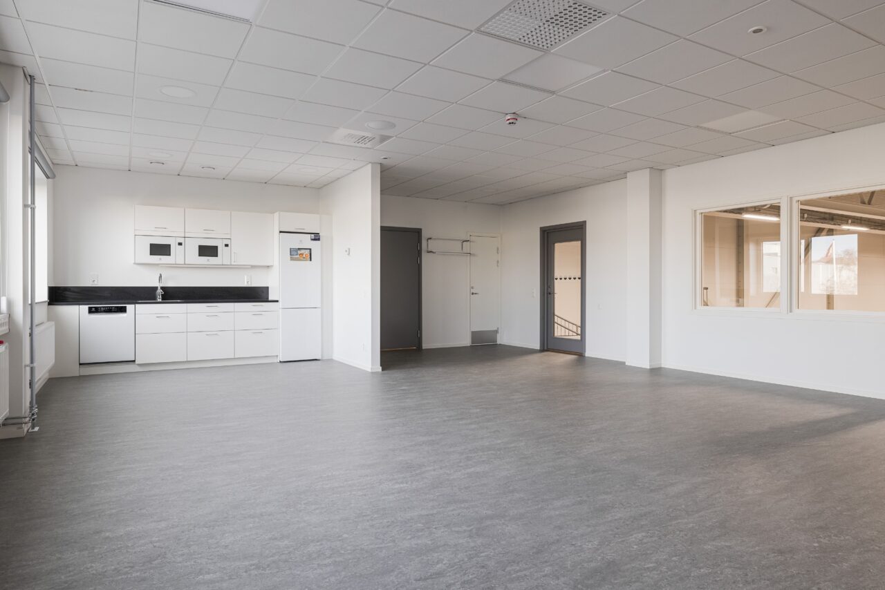 Omöblerat, modernt rum med grått golv, vita väggar, pentry till vänster, stora innerfönster och en stängd grå dörr till höger.