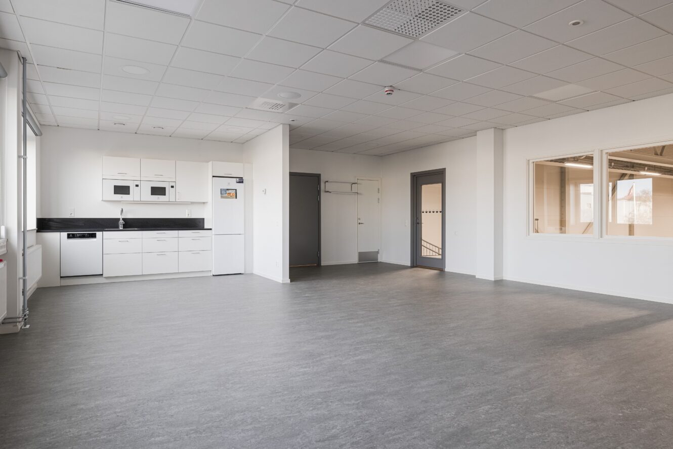 Omöblerat, modernt rum med grått golv, vita väggar, pentry till vänster, stora innerfönster och en stängd grå dörr till höger.