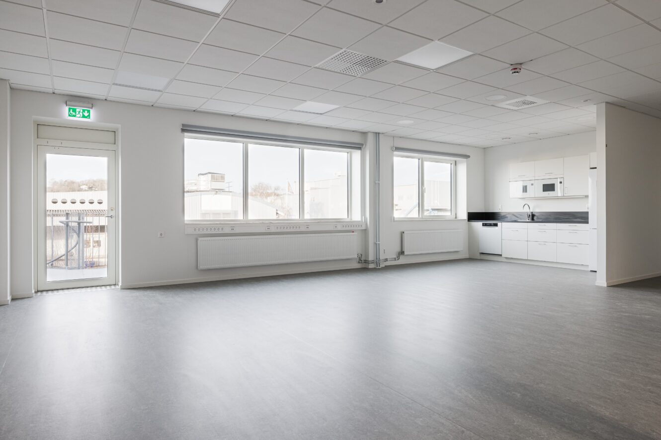 Rymligt, omöblerat rum med stora fönster, ett pentry med vita skåp och vitvaror, grått golv och en dörr som leder ut.