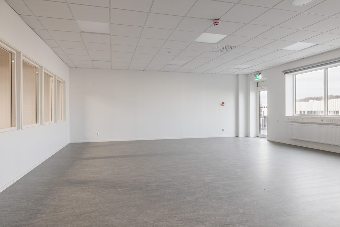 Ett tomt, omöblerat rum med vita väggar, grått golv, takplattor, stora fönster och en glasdörr som leder ut.