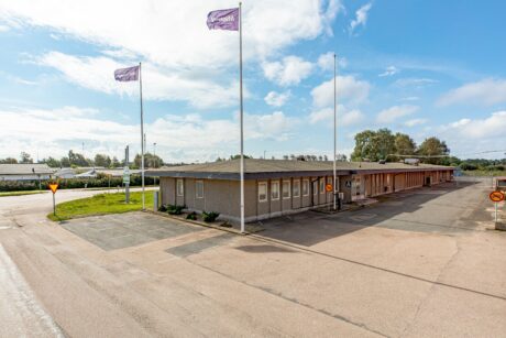 Envåningsbyggnad med flaggstänger framför, omgiven av en asfalterad yta och låg grönska, under en delvis molnig himmel.