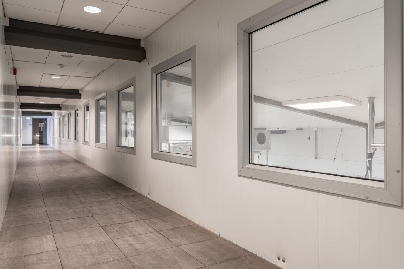 En lång, tom korridor med klinkergolv och stora fönster till höger, med utsikt över en ljus industri- eller lagerlokal.