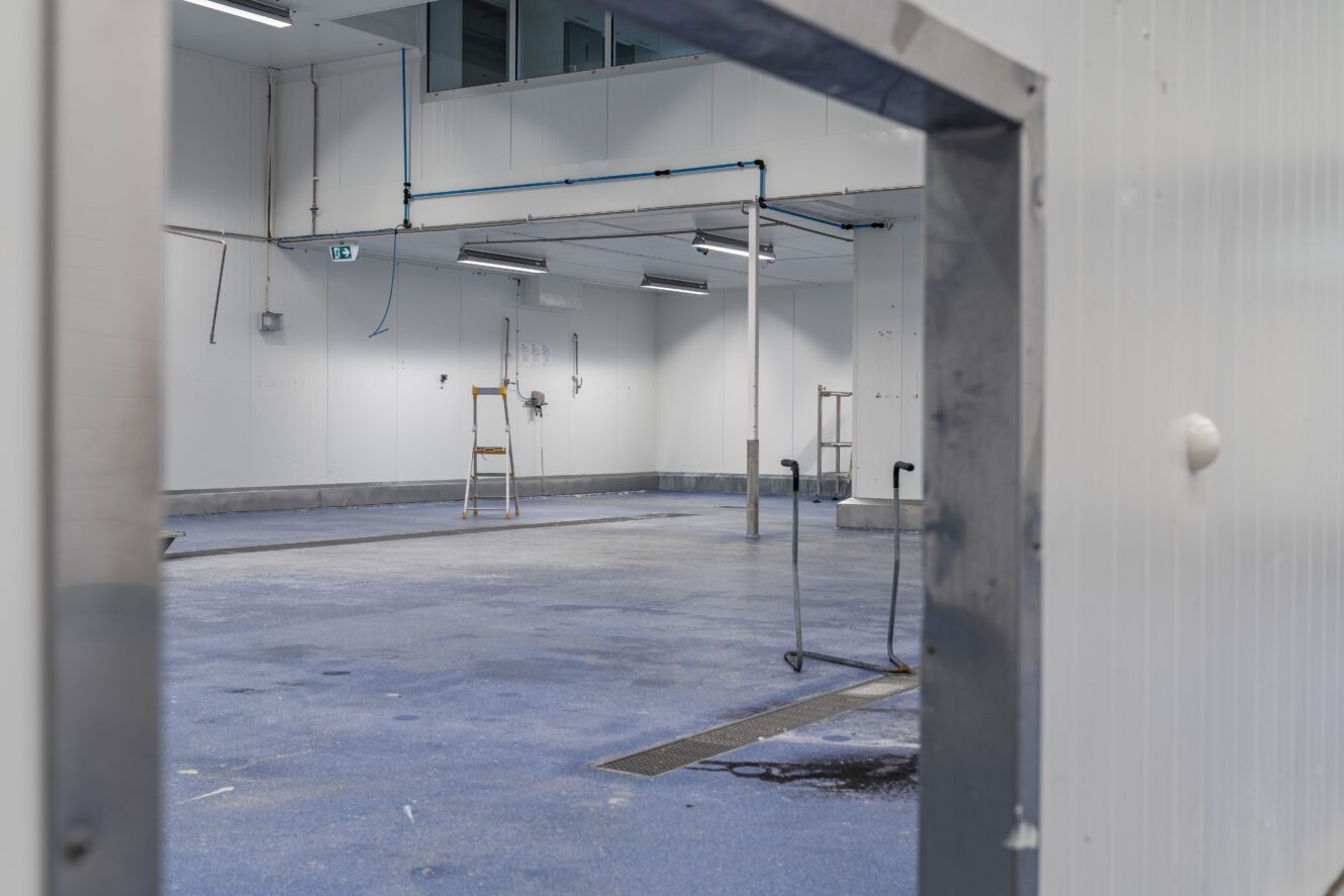 En tom industrilokal med blått golv, vita väggar, rörledningar, en trappstege och en dörrkarm i metall i förgrunden.
