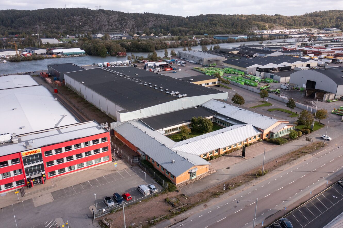 Flygfoto över ett industriområde med lagerlokaler, en röd DHL-byggnad, vägar, parkerade bilar och gröna containrar nära en vattensamling och skogsklädda kullar.