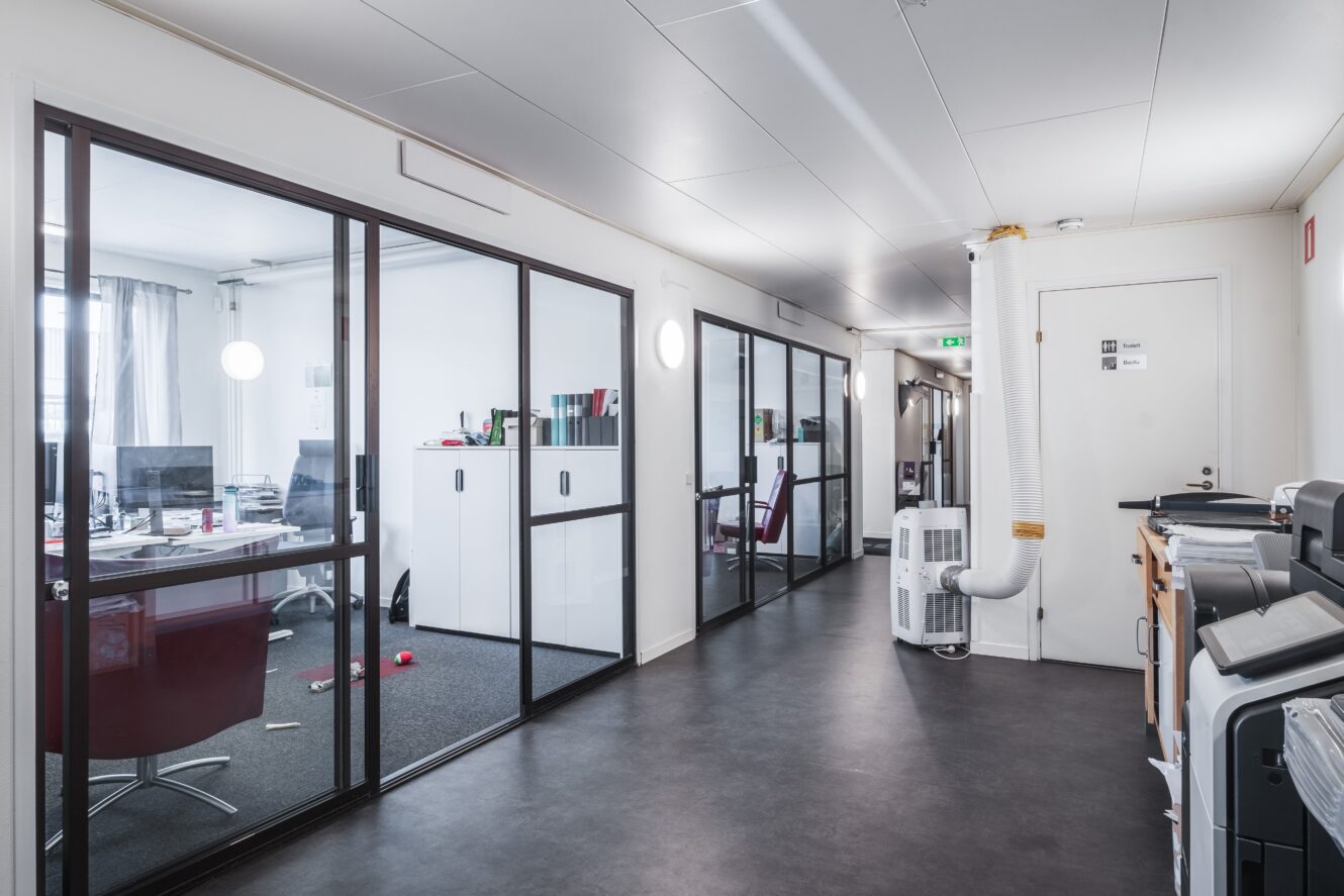 Modern kontorskorridor med rum med glasväggar till vänster, kontorsutrustning till höger och ett portabelt luftkonditioneringsaggregat i slutet av korridoren.