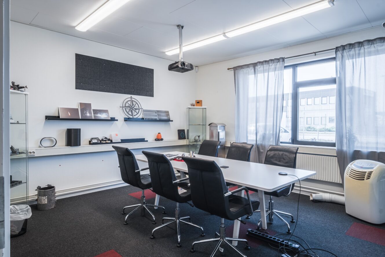 Modernt konferensrum med ett rektangulärt bord, sex stolar, en projektor, vitrinskåp, ett fönster med skira gardiner och olika kontorsartiklar på hyllor.