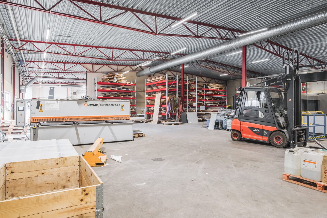 Inredning i ett industrilager med metallställ, en gaffeltruck, en skärmaskin och olika material och utrustning som är organiserade i hela utrymmet.