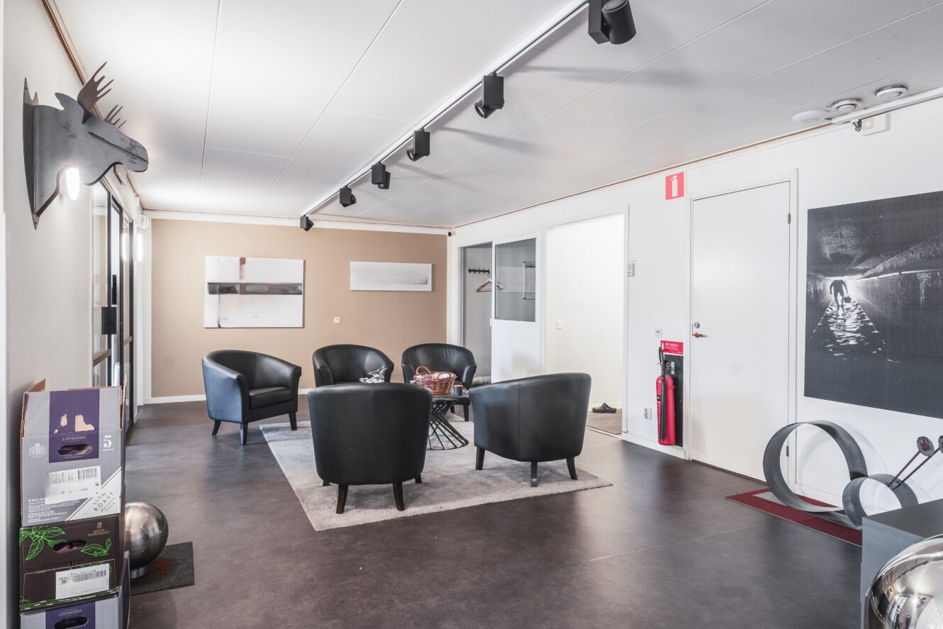 Modernt väntrum med fyra svarta fåtöljer runt ett soffbord, väggkonst, klädhängare och brandutrustning nära dörren.