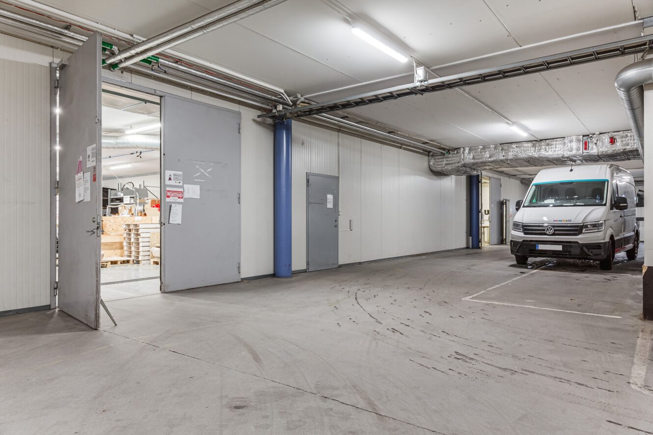 En vit skåpbil står parkerad i ett industriellt garage inomhus nära öppna dörrar som leder till en arbetsplats med staplade pallar och utrustning. Rör i taket och lysrör är synliga.