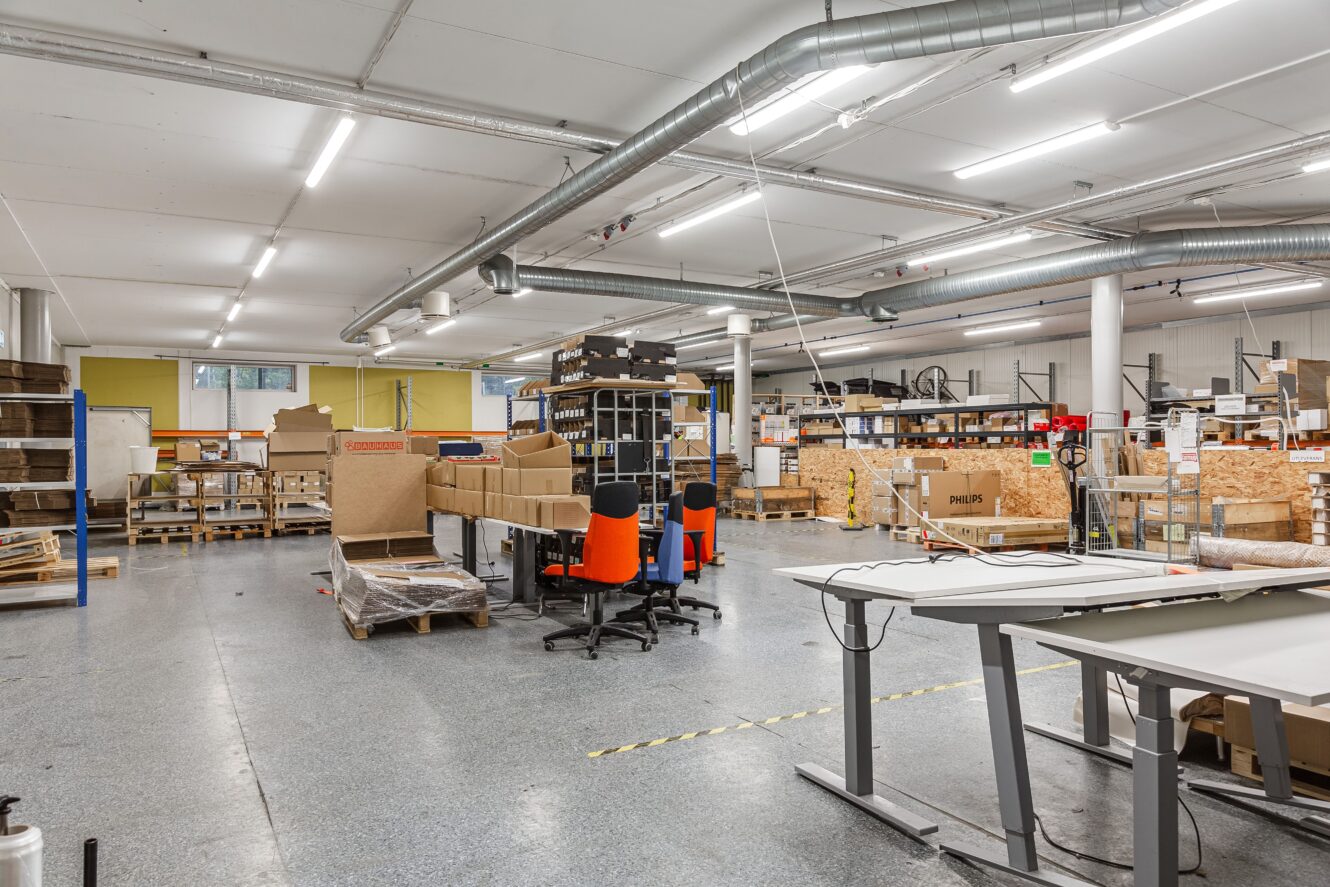 En rymlig lagerlokal med hyllor fyllda med lådor och material, utspridda arbetsstationer, orangefärgade stolar och industriell belysning ovanför.