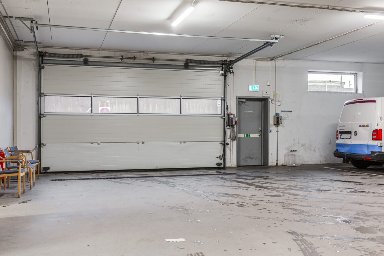 Ett rent, tomt garage med en stor takskjutport, flera stolar uppradade mot väggen och en vit skåpbil parkerad nära en grå utgångsdörr.