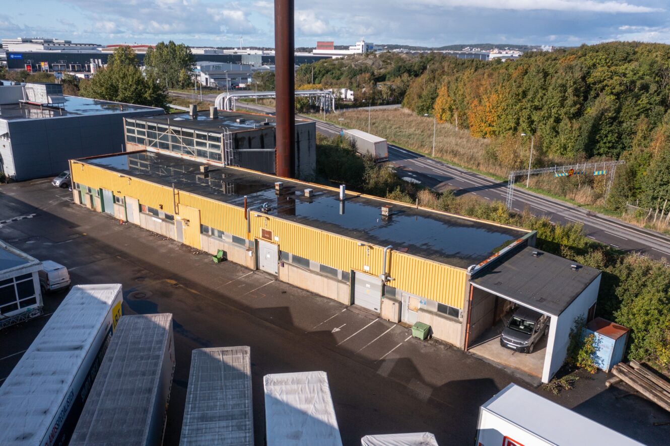 Flygfoto över en lång, gul industribyggnad med platt tak, omgiven av lastbilar och grönska, intill en väg och träd.