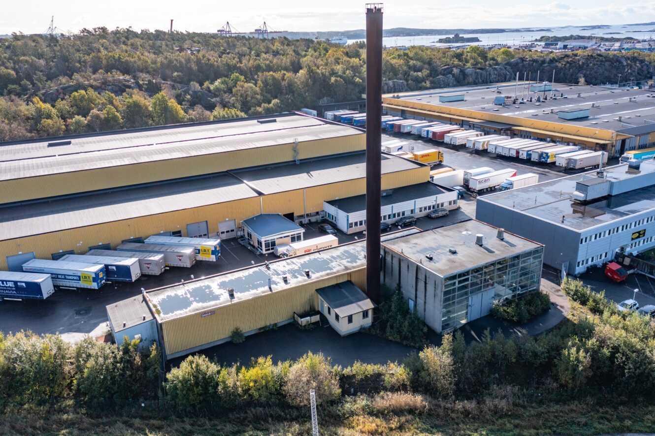 Flygfoto över en stor industrianläggning med gula lagerbyggnader, en hög skorsten, parkerade lastbilar och omgivande grönska.